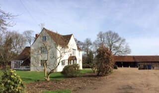 A large white farmhouse with a sloping red roof sits among green grass and trees, with a dirt drive and a barn or shed in the background under a clear sky.
