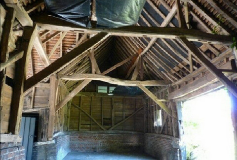 Interior of an old wooden barn with exposed beams, brick lower walls, and a high, slanted roof. Sunlight enters from an open doorway on the right, illuminating the rustic structure.