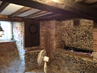 A rustic room with exposed brick and stone walls, wooden ceiling beams, a dartboard mounted on the wall, and a white oscillating fan on the floor. Light enters through a window on the left.