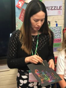 A woman with long brown hair holds a scratch art drawing and uses a stylus to reveal colourful patterns. She stands in a classroom near a child, with educational posters and children's artwork on the wall behind her.
