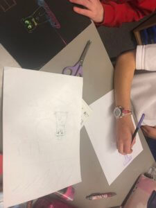 Two children are sitting at a desk drawing on white paper with pencils. A pair of purple scissors and coloured pencils are on the table. One child wears a red top and a watch, while sketches and art supplies are visible.
