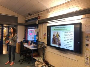 A woman stands in a classroom giving a presentation. A large screen displays a slide titled Story Book Illustration: An Anglo-Saxon Adventure with text and an illustration of Anglo-Saxon figures.