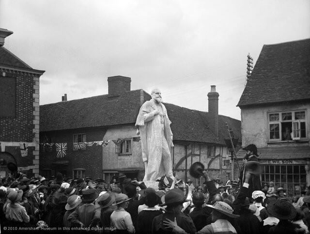 A crowd gathers around a large statue of a bearded man in a town square, with people in old-fashioned clothing. Historic buildings and bunting flags evoke the charm of Amersham film locations.