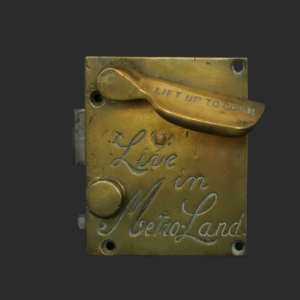 A brass mechanical latch with the engraved words Live in Metro-Land and a lever handle labelled LIFT UP TO OPEN, set against a dark background.