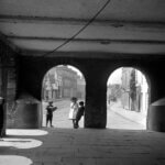Black-and-white photo of five people standing and talking under a brick archway that opens onto a quiet street lined with old buildings. Sunlight streams in through the arches, casting shadows on the stone floor.