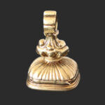 A gold fob seal with an ornate, curved handle and a ridged, cushion-shaped base, set against a plain dark background.