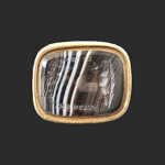 A rectangular, dark stone with white streaks, set in a simple gold bezel, displayed against a solid dark background.