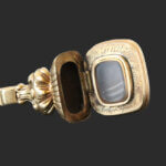 A gold locket with ornate detailing, shown open to reveal an empty compartment and an inscribed message along the inner rim. The locket is displayed against a plain dark background.