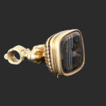 A gold ring with an ornate, square-faced black gemstone featuring an engraved design, displayed against a plain dark background.