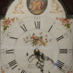An antique clock face with Roman numerals, ornate hands, floral decorations, a painted bird in the centre, and an illustration of a seated woman at the top, surrounded by gold and rose details.