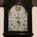 An antique grandfather clock with a dark wooden case, a decorative face with Roman numerals, floral patterns, and a small painting of a seated figure at the top. The clock shows the time as 12:27.