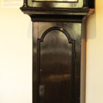 A tall, dark wooden grandfather clock with a decorated dial showing Roman numerals, ornate metal hands, and a painted scene above the clock face, standing against a light-coloured wall.
