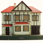 A vintage, two-storey toy dolls' house with a red roof, pale blue walls, black trim, multiple windows, a front door with a small staircase, and a painted garden by the entrance.