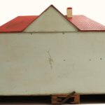 A simple model house with a red tiled roof, white walls, and a chimney, elevated on wooden pallets. The front wall is plain with minor marks and no windows or doors visible.