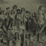 A vintage photograph shows a large group of people in elaborate costumes, including various hats, crowns, and theatrical outfits, posing outdoors in a staged, formal arrangement.