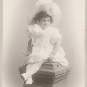 A young girl in a frilly white dress and large hat sits on a stone pedestal, posing for a vintage studio portrait. The photo has a faded sepia tone and ornate text at the bottom with the photographer’s name and address.