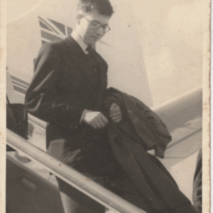 A young man in a suit and glasses descends aeroplane stairs, holding a coat. The image is black and white and appears vintage; part of an aircraft tail with a painted design is visible in the background.