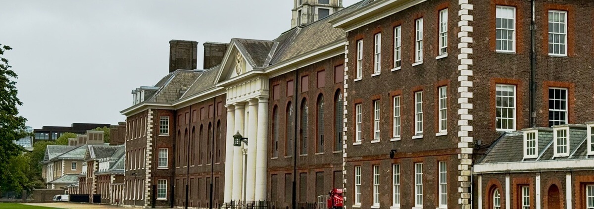 A long, historic red-brick building with white columns and sash windows, featuring a small clock tower on the roof. All aboard for Amersham—a person in red stands near the entrance on a cloudy day.