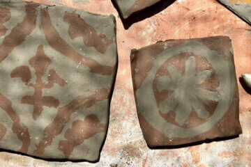 Two square clay tiles with brown and grey designs, one featuring a floral pattern and the other a circular motif, are placed on a textured surface next to two yellow sculpting tools.