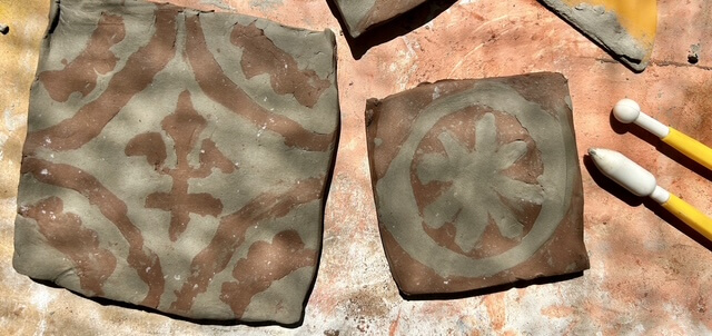 Two square clay tiles with brown and grey designs, one featuring a floral pattern and the other a circular motif, are placed on a textured surface next to two yellow sculpting tools.