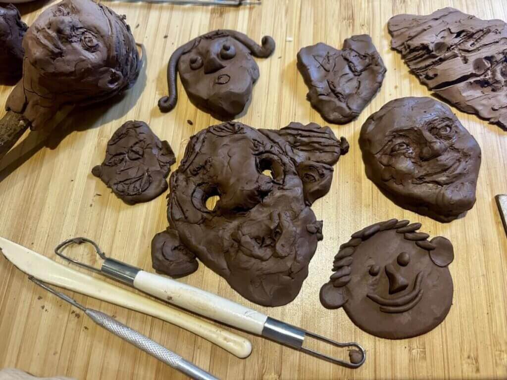 Several clay faces and sculpting tools are arranged on a wooden surface, showcasing different facial expressions and styles, some detailed and others more abstract or rough.