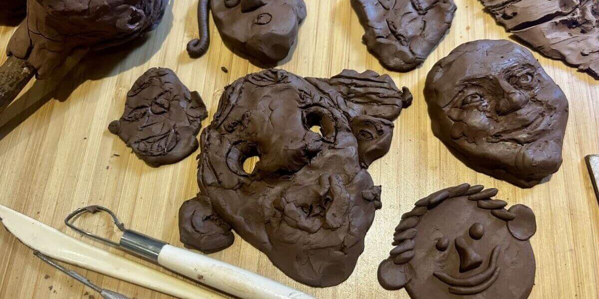 Several clay faces and sculpting tools are arranged on a wooden surface, showcasing different facial expressions and styles, some detailed and others more abstract or rough.
