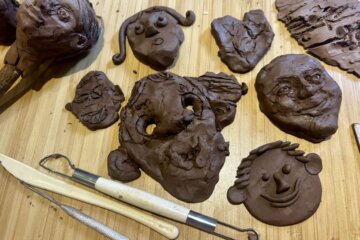 Several clay faces and sculpting tools are arranged on a wooden surface, showcasing different facial expressions and styles, some detailed and others more abstract or rough.