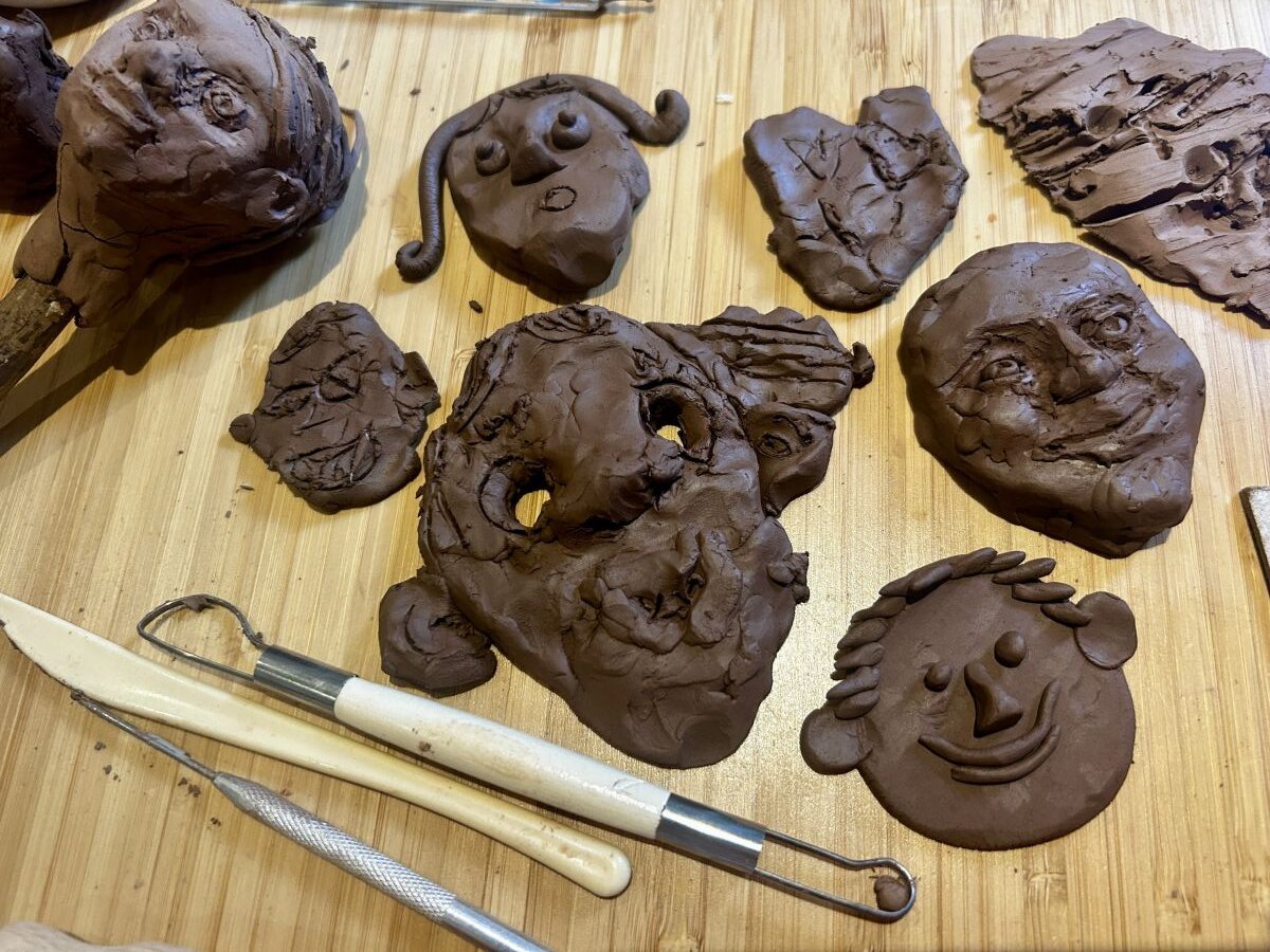 Several clay faces and sculpting tools are arranged on a wooden surface, showcasing different facial expressions and styles, some detailed and others more abstract or rough.