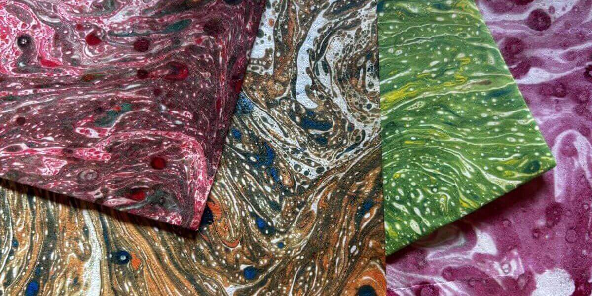 Four sheets of marbled paper overlap, each with swirling patterns in red, brown, green, and pink tones. Small dots of contrasting colours are scattered throughout the designs.