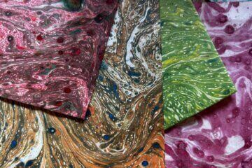 Four sheets of marbled paper overlap, each with swirling patterns in red, brown, green, and pink tones. Small dots of contrasting colours are scattered throughout the designs.
