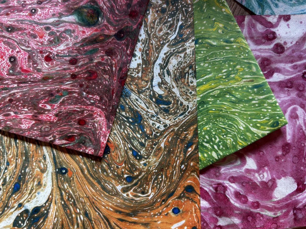 Four sheets of marbled paper overlap, each with swirling patterns in red, brown, green, and pink tones. Small dots of contrasting colours are scattered throughout the designs.