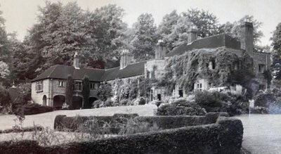 Black and white photo of a large, historic mansion surrounded by trees and hedges. The building is covered in ivy, with several chimneys and arched windows. The foreground shows neatly trimmed bushes and a garden.