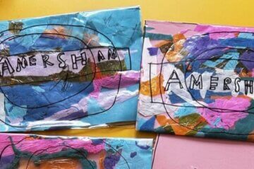 Three colourful collages with overlapping paint and paper feature the word AMERSHAM outlined in black. The backgrounds are blue, pink, and multicoloured, and the artworks are spread out on a table.