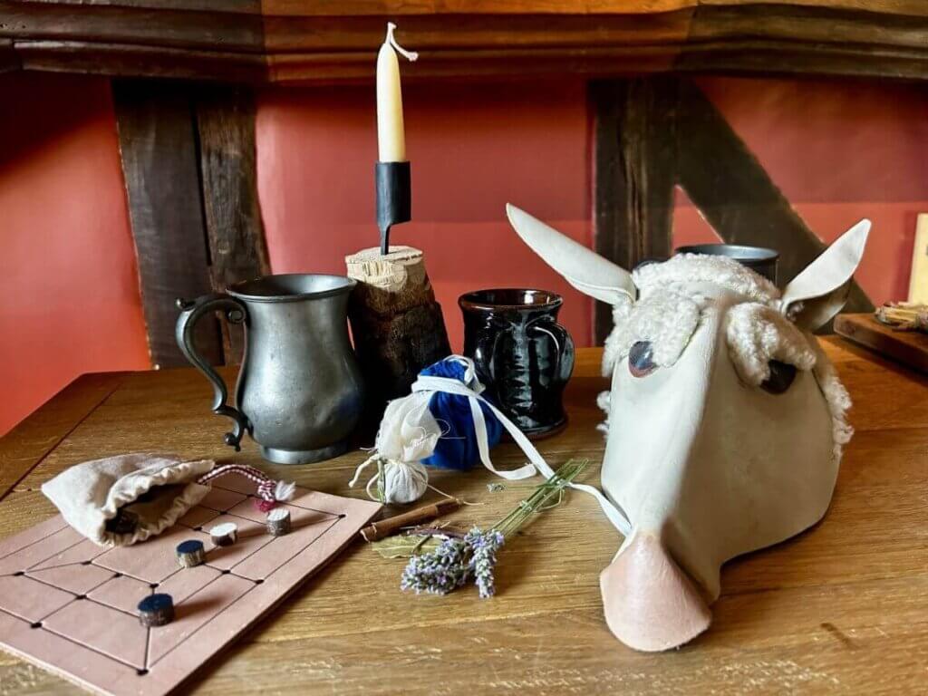 A wooden table with a metal mug, black cup, candle, sheep mask, dried lavender, cloth bag, pouch, and a simple board game with black and white pieces sets the scene in a rustic Tudor Crafts workshop filled with red walls and dark wood beams.
