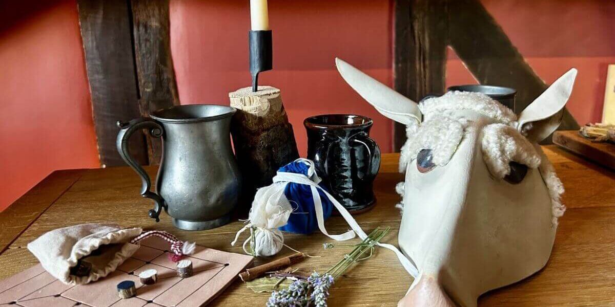 A wooden table with a metal mug, black cup, candle, sheep mask, dried lavender, cloth bag, pouch, and a simple board game with black and white pieces sets the scene in a rustic Tudor Crafts workshop filled with red walls and dark wood beams.