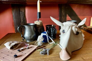 A wooden table with a metal mug, black cup, candle, sheep mask, dried lavender, cloth bag, pouch, and a simple board game with black and white pieces sets the scene in a rustic Tudor Crafts workshop filled with red walls and dark wood beams.