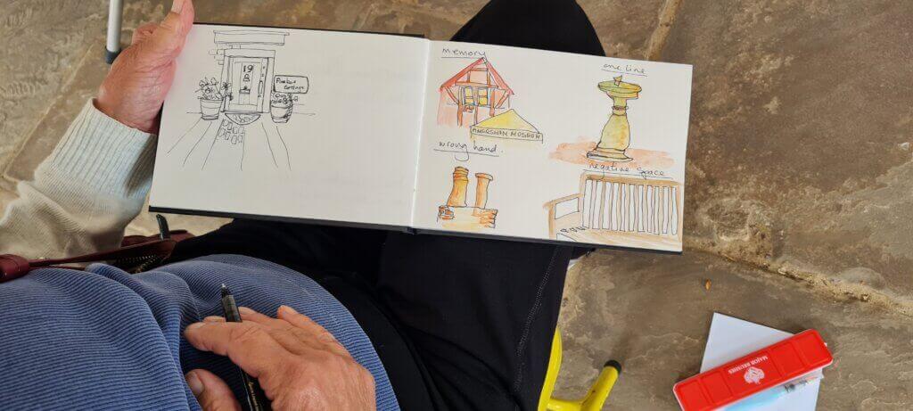 A person sketching in a notebook displays drawings of a doorway, buildings, a statue, and a railing, some in colour. The person holds a pen and sits on a stone floor with a red pencil case nearby.