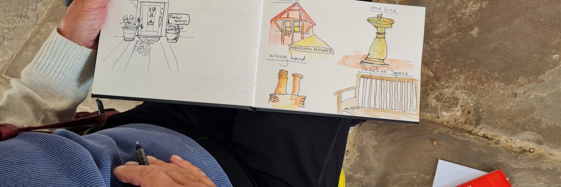 A person sketching in a notebook displays drawings of a doorway, buildings, a statue, and a railing, some in colour. The person holds a pen and sits on a stone floor with a red pencil case nearby.