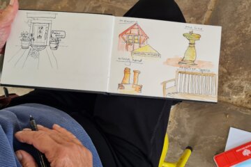 A person sketching in a notebook displays drawings of a doorway, buildings, a statue, and a railing, some in colour. The person holds a pen and sits on a stone floor with a red pencil case nearby.
