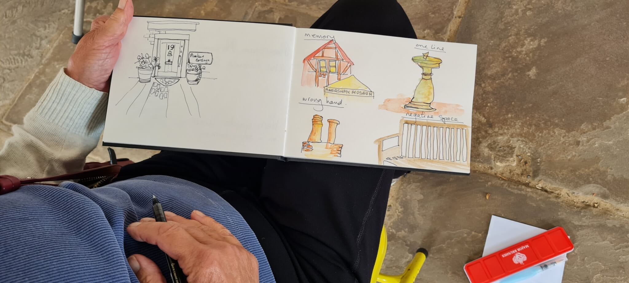 A person sketching in a notebook displays drawings of a doorway, buildings, a statue, and a railing, some in colour. The person holds a pen and sits on a stone floor with a red pencil case nearby.