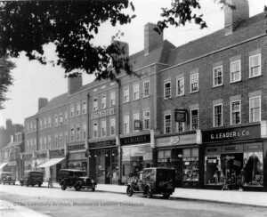 Black and white photo of a row of shops on a city street with parked vintage cars in front. The buildings have multiple storeys and large windows, and the street is quiet with few people visible.