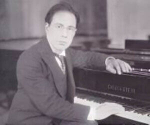 A man in a suit sits at a grand piano with one hand on the keys and the other resting on the piano, looking towards the camera in a black and white photo, evoking the atmosphere of an intimate concert talk with Francesco Ticciati.