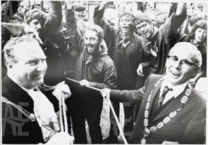 Black-and-white photograph of two officials in ceremonial chains and cheering crowds.