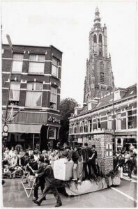 Black-and-white historical photograph shows a crowded street parade and a decorated float