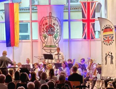 Photograph shows a brass band performing onstage before a seated audience with the German flag, the Union Jack flag and the banner of the Amersham Bensheim Society above