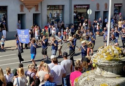 Photograph showing a lively street parade with a marching brass band performing before a large crowd