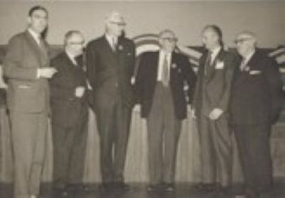 Black-and-white group photograph shows six formally dressed men standing on a stage at a civic event