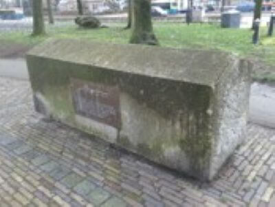 Photograph of a weathered rectangular stone monument with moss-covered surfaces and a bronze plaque set into its front