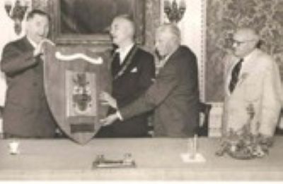 Black-and-white photograph shows four men at a formal presentation, holding a large decorative shield or plaque