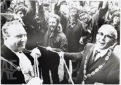 Black-and-white photograph of two officials in ceremonial chains and cheering crowds